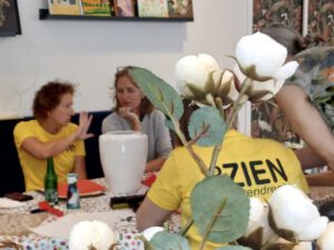 Foto genomen vanachter bloemen, Esther en Marjolein zijn bezig met een vaas, Dennes vanop de rug gezien.